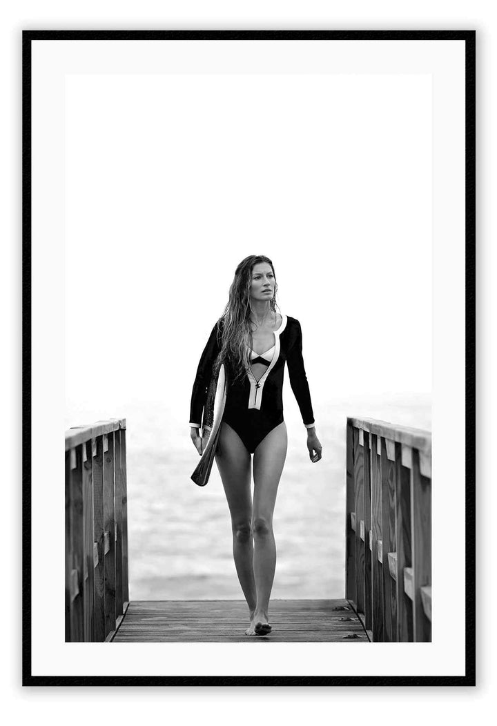 A black and white fashion wall art with Brazilian model Gisele Bundchen holding a surf board walking on timber wharf.