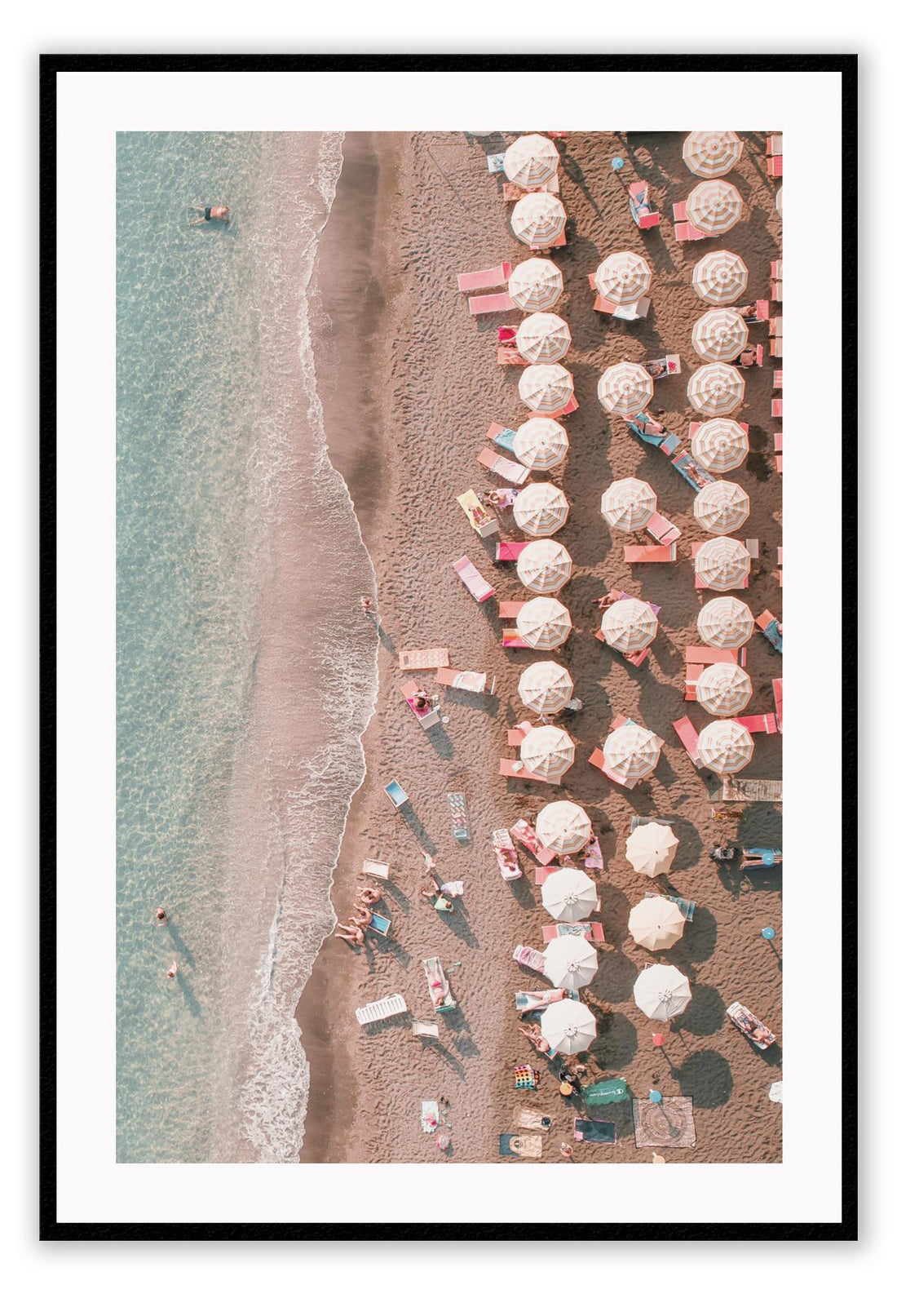 Coastal style photography beach print with pink umbrellas and chairs on the sand in front of the water from a birds eye view.